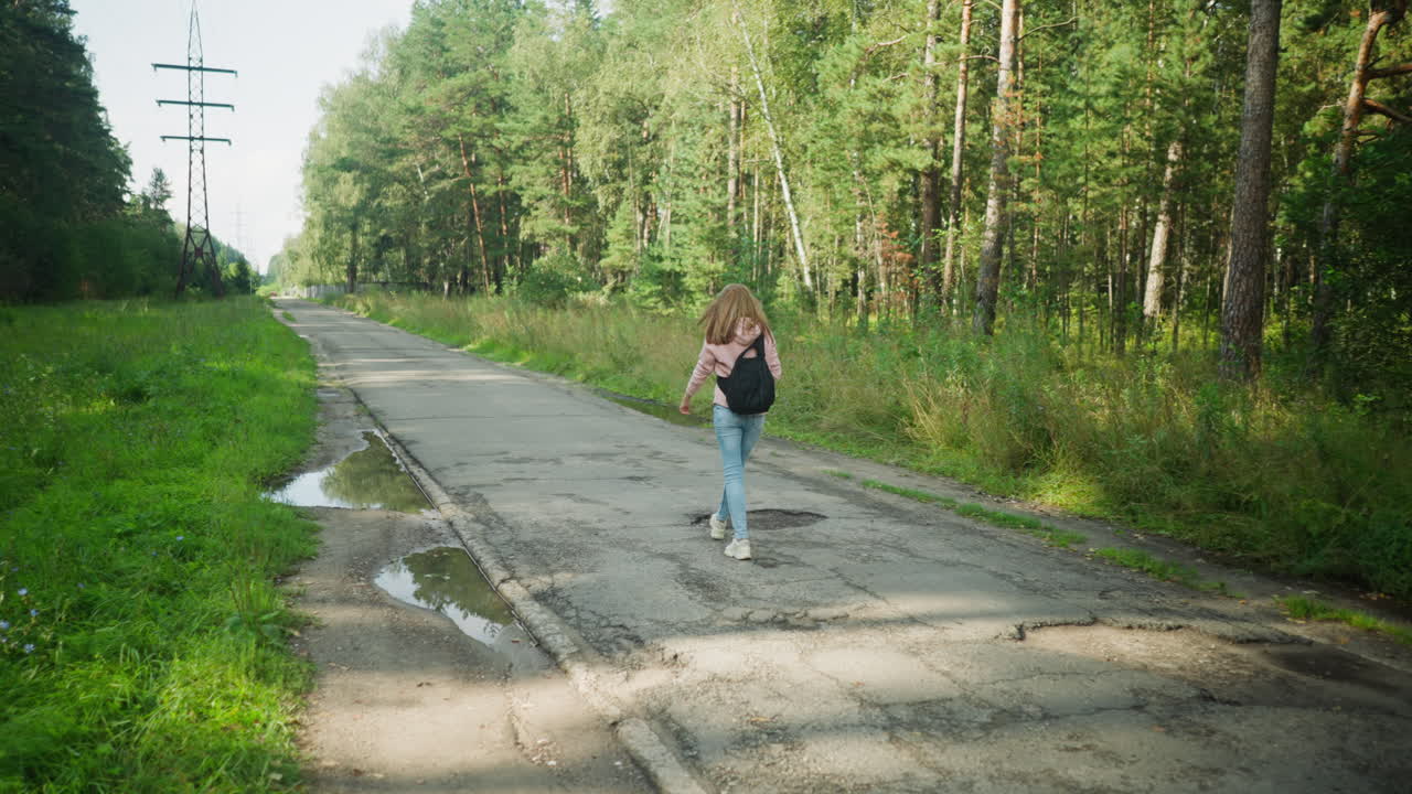 Back view of tourist walking alone with head bowed on cracked tarred road surrounded by forest and overhead power lines, casting soft shadows across puddles and greenery under bright
