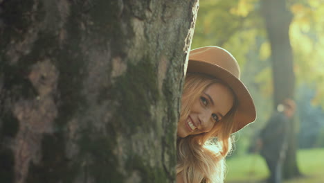 joven rubia caucásica con sombrero, sosteniendo una hoja amarilla y mirando la cámara detrás de un árbol en el parque en otoño