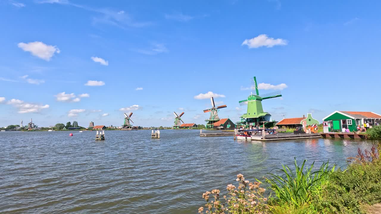 Wide shot of Dutch windmills, canal, and green houses under bright daylight, camera remains steady