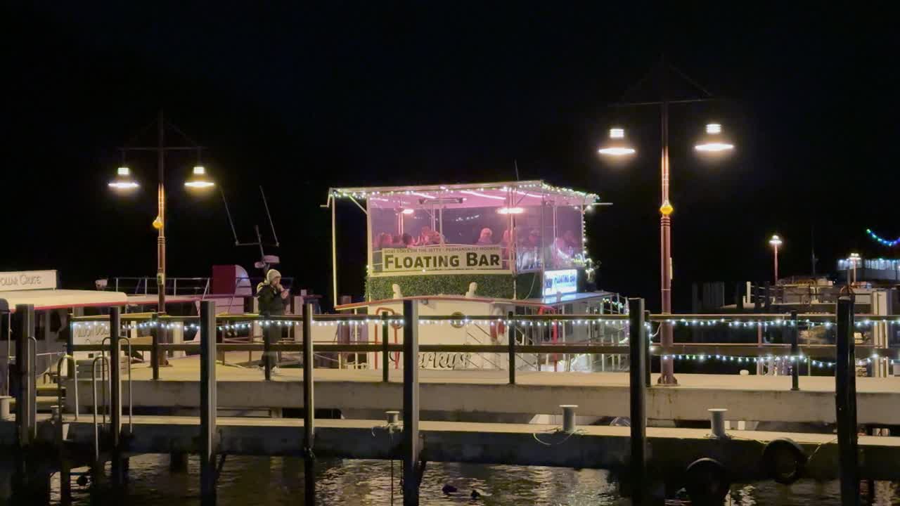 A floating bar illuminated at night on a calm waterfront in Queenstown, New Zealand, with vibrant lighting and serene ambiance