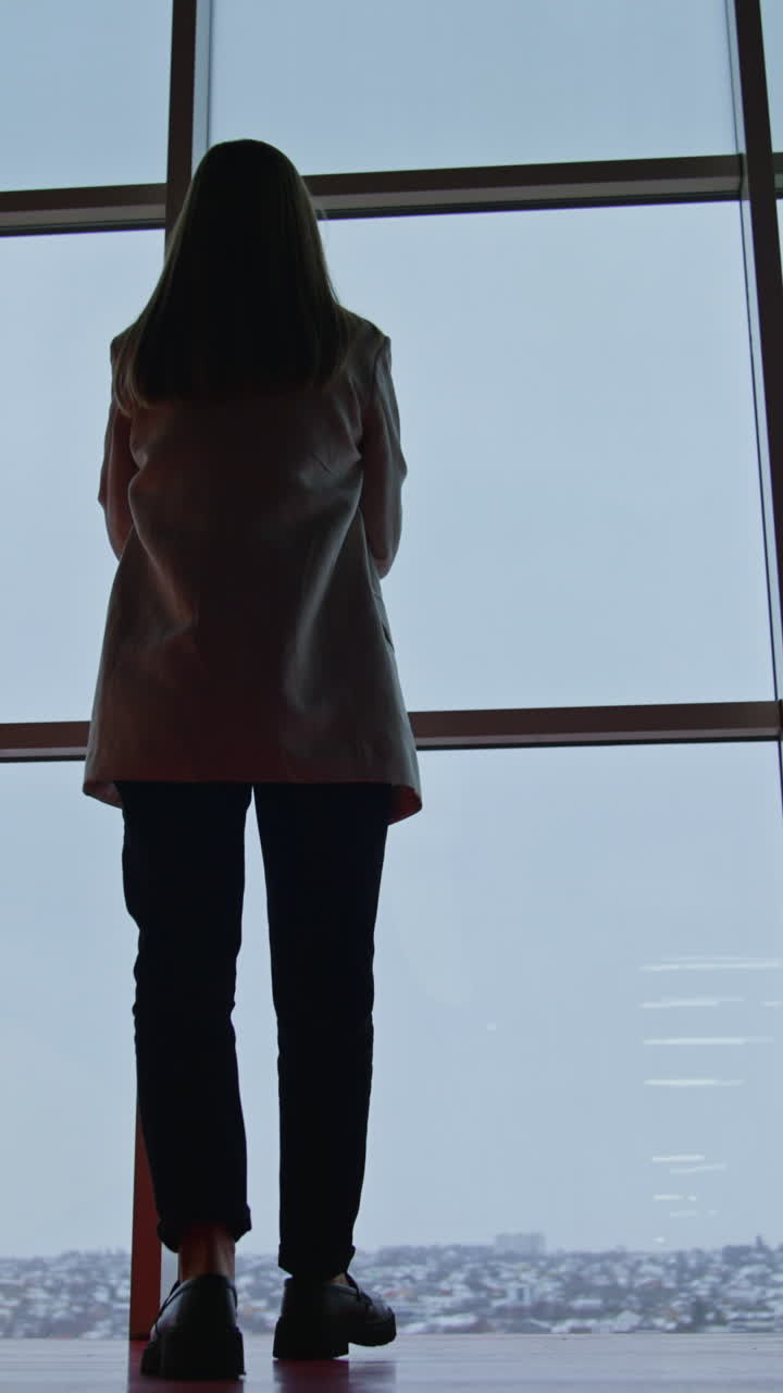 Woman in suit comes up to the big panoramic windows. Thoughtful young lady standing and watching outside. Low angle view. Vertical video