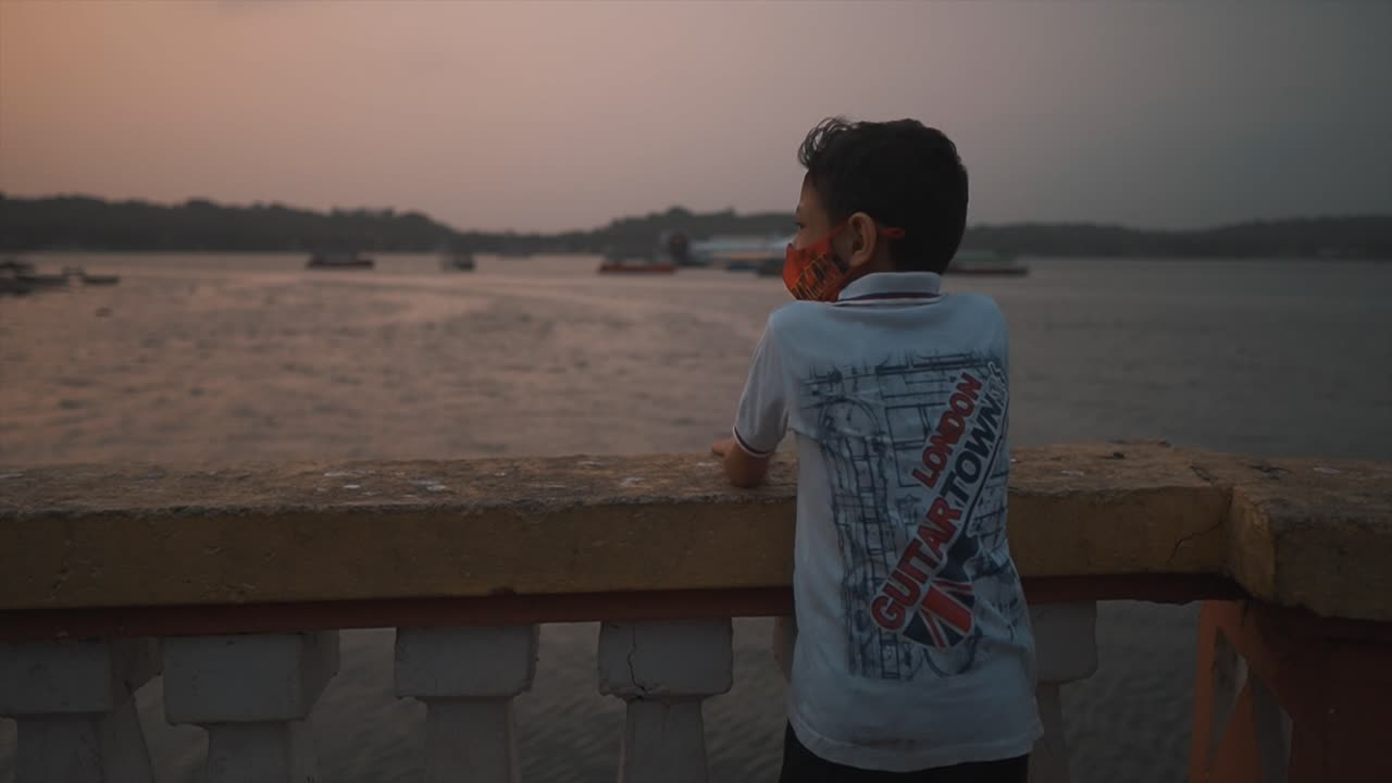 un primer plano de un joven entusiasta que señala diferentes barcos parados detrás de una balaustrada de hormigón que domina el hermoso río mandovi al atardecer, goa, india