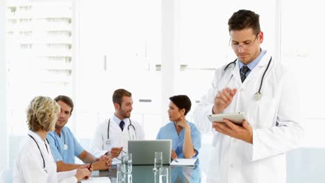 Thoughtful-doctor-using-tablet-with-staff-talking-behind-him-