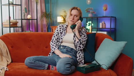 Smiling-young-woman-making-wired-telephone-call-conversation-with-friends-sitting-on-home-couch