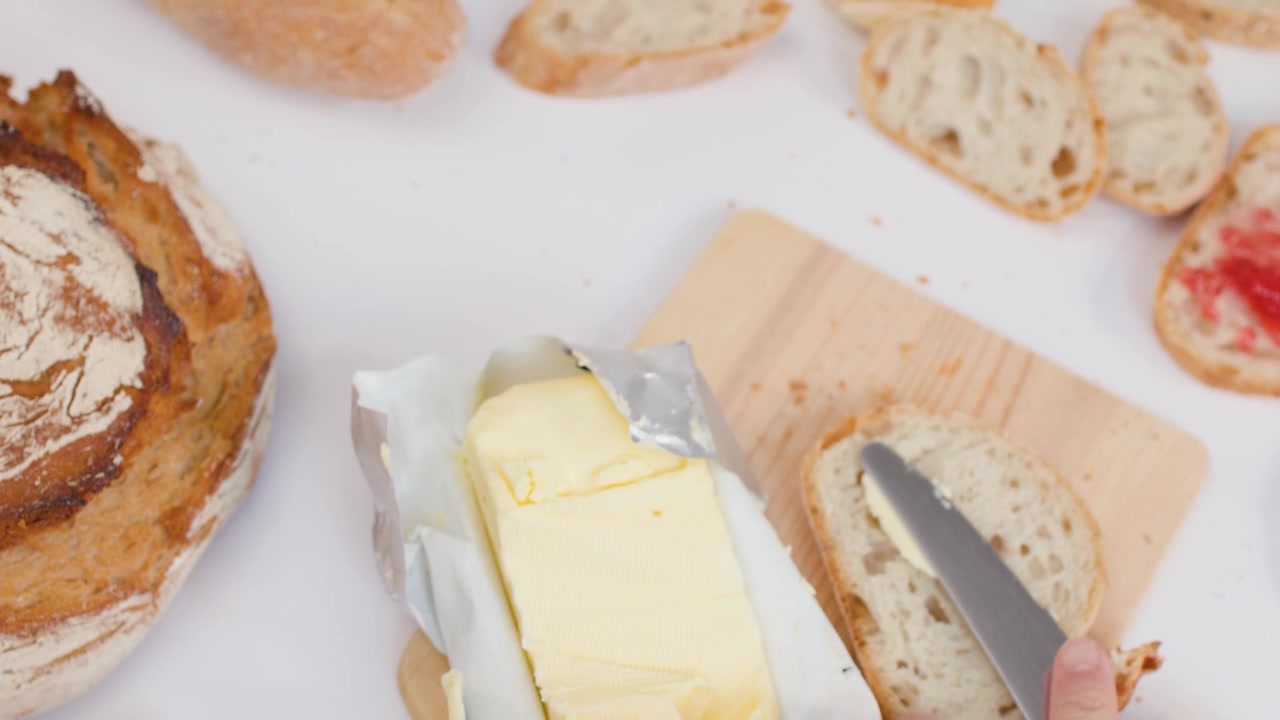 Ripresa dall'alto di mani che tagliano burro fresco e lo spalmano su fette di pane croccante al lievito madre.