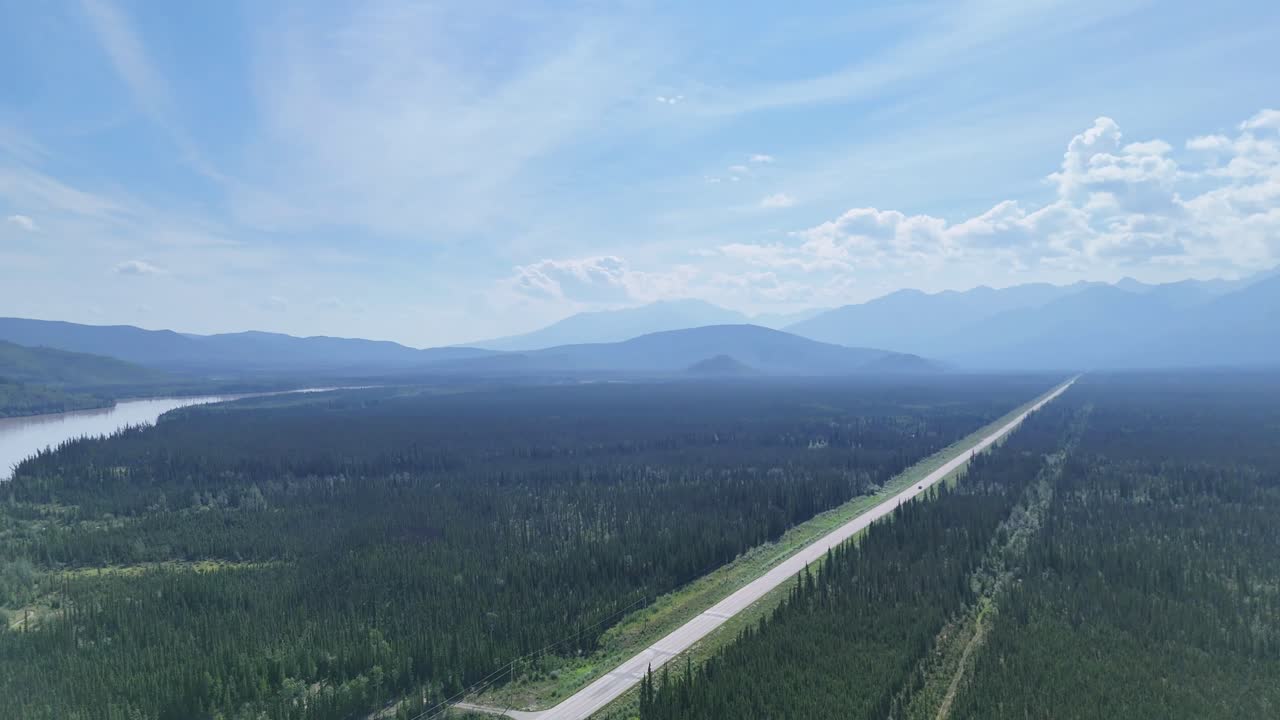 Stunning aerial footage of a car driving down a long highway cutting through a dense forest with majestic mountains in the distance under a bright blue sky in Alaska