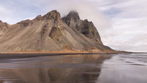 vestrahorn’s jagged peaks shrouded in clouds, mirrored on the black sands of stokksnes. iceland