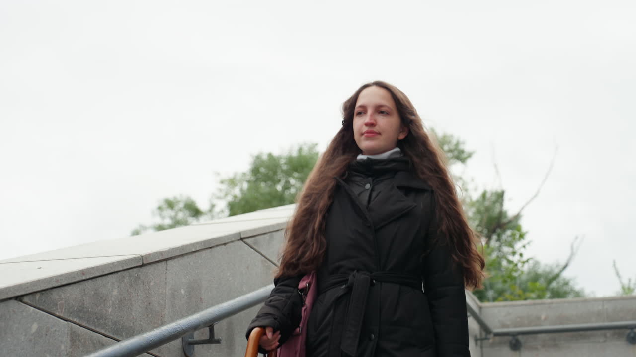 Donna in città con ombrello. Donna con cappotto nero che scende le scale. Persona che passeggia sui gradini in ambiente urbano. Donna con ombrello e cappotto che percorre le scale della città durante una giornata nuvolosa