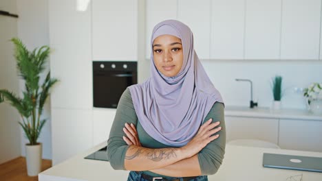 modern young tattooed arab woman in a lilac hijab stands in the kitchen with her arms folded. muslim wife with beautiful face with eyelash extensions and piercings. womens rights in the middle east