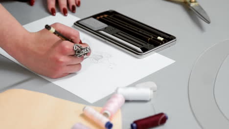 Designer-drawing-sketches-for-clothes.-Hipster-designer-working-from-home,-sketching-a-fashion-project.-Female-artist's-hand-sketching-with-a-pencil-on-a-light-day.-Closeup.