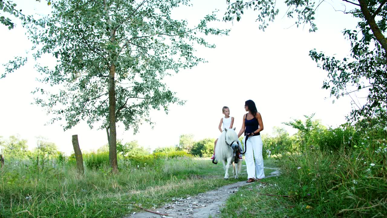 A woman and a boy are walking around the field, son is riding a pony, mother is holding a pony for a bridle. Cheerful, happy family vacation. Outdoors, in summer, near the forest