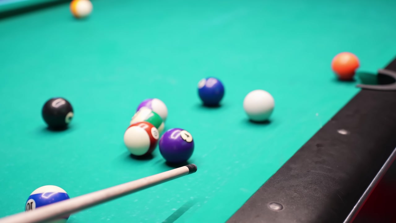 Pool cue ball strikes a tight cluster of colorful balls on green felt, creating a dynamic collision and scatter near the rail in a cinematic bar billiards scene.