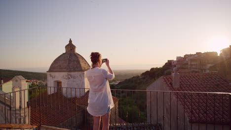 happy woman using smartphone taking photo of beautiful sunset enjoying sharing summer vacation travel experience on balcony