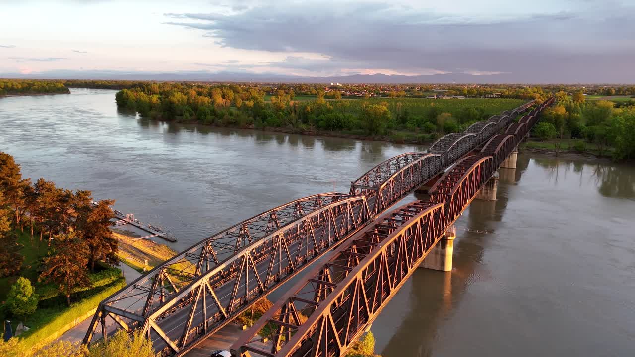 Al tramonto, i pendolari attraversano il vecchio ponte di ferro che attraversa il fiume Po, segnando il confine tra Lombardia ed Emilia-Romagna, dove ferrovia e strada collegano Cremona a città e regioni vicine.