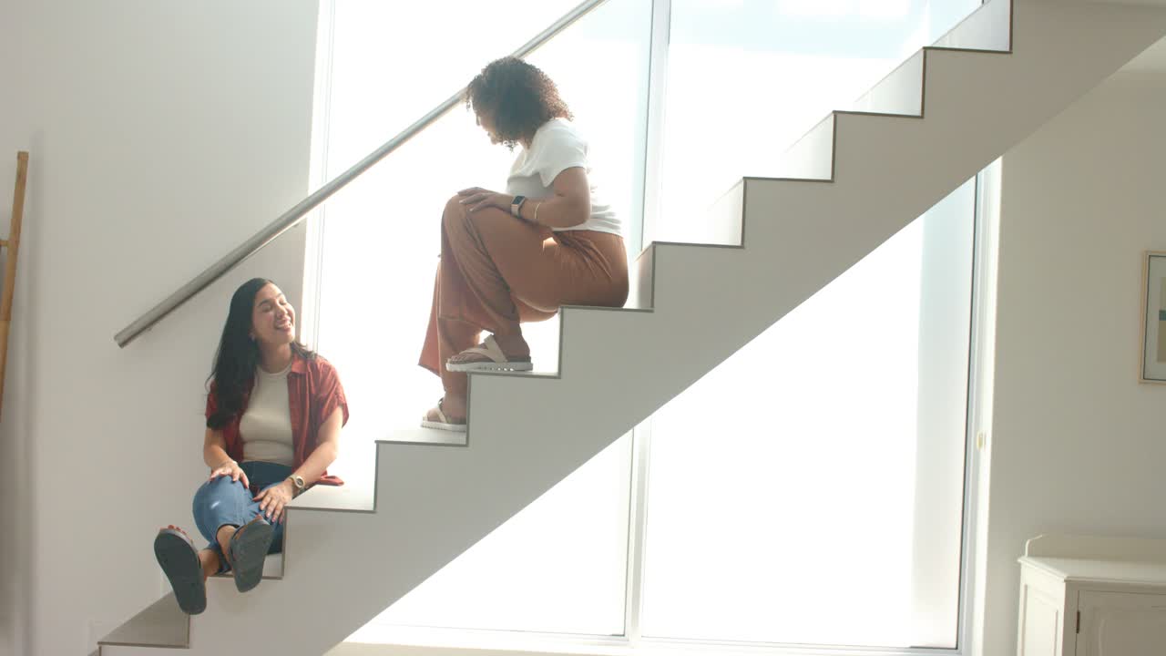 Twee vrouwen in casual kleding kletsen, zittend op een open trap bij het raam, met metalen leuning