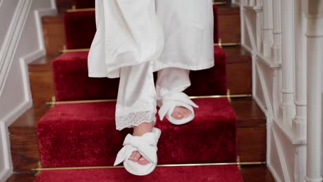 bride in white robe walks down red carpeted stairs holding bouquet in soft natural light