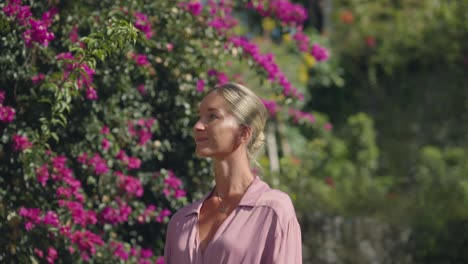 encantadora mujer rubia vestida de rosa caminando junto a flores tropicales de color púrpura buganvillas
