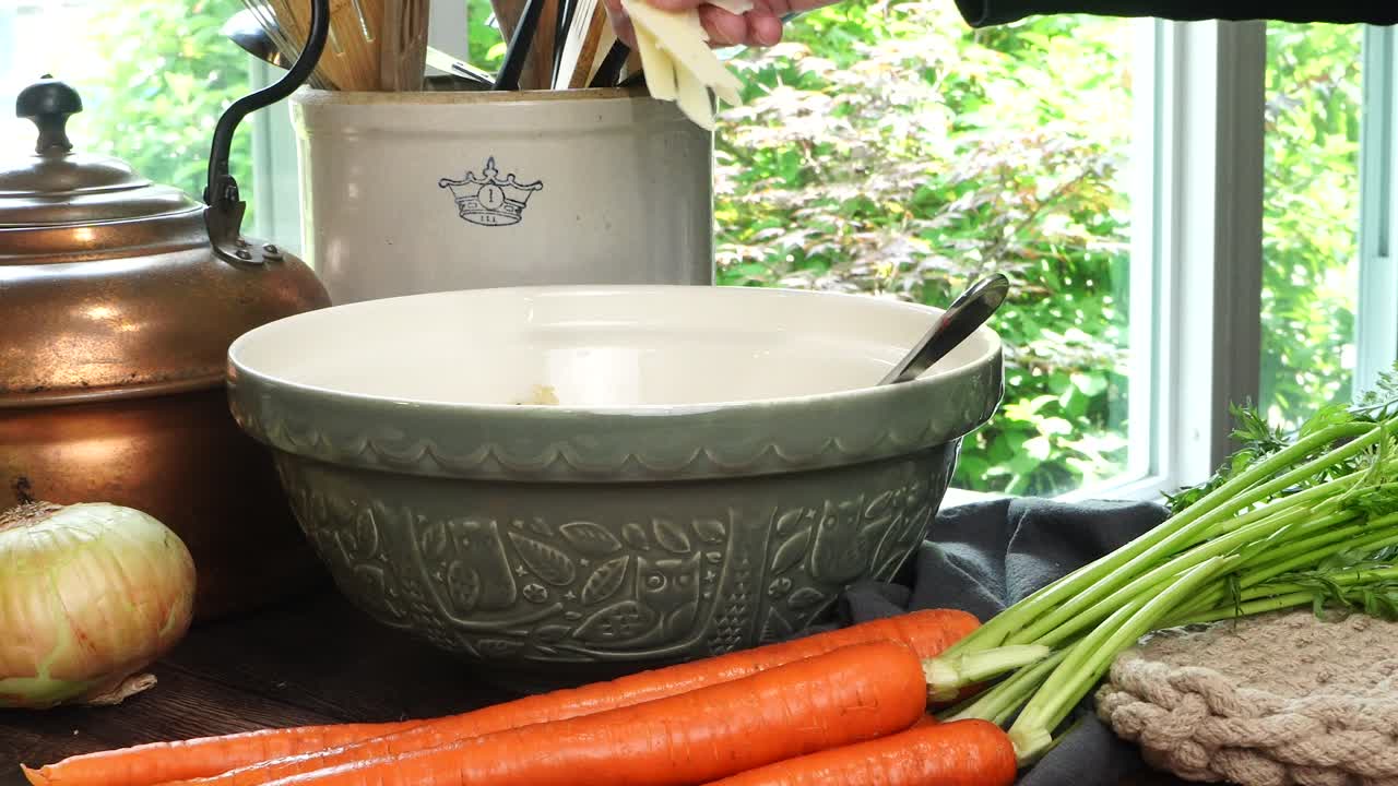 Making meatballs for Italian Wedding Soup. Adding parmesan cheese with hands into a big green ceramic bowl on a counter in the kitchen. Fresh vegetables like carrots and onions on the counter.