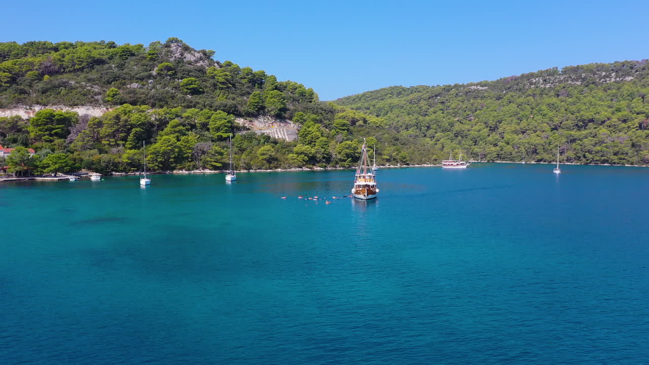 Sailboat moored in beautiful blue water, tourists swimming in sea, aerial dolly