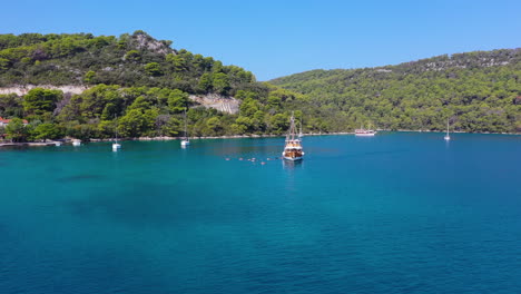 Sailboat-moored-in-beautiful-blue-water,-tourists-swimming-in-sea,-aerial-dolly