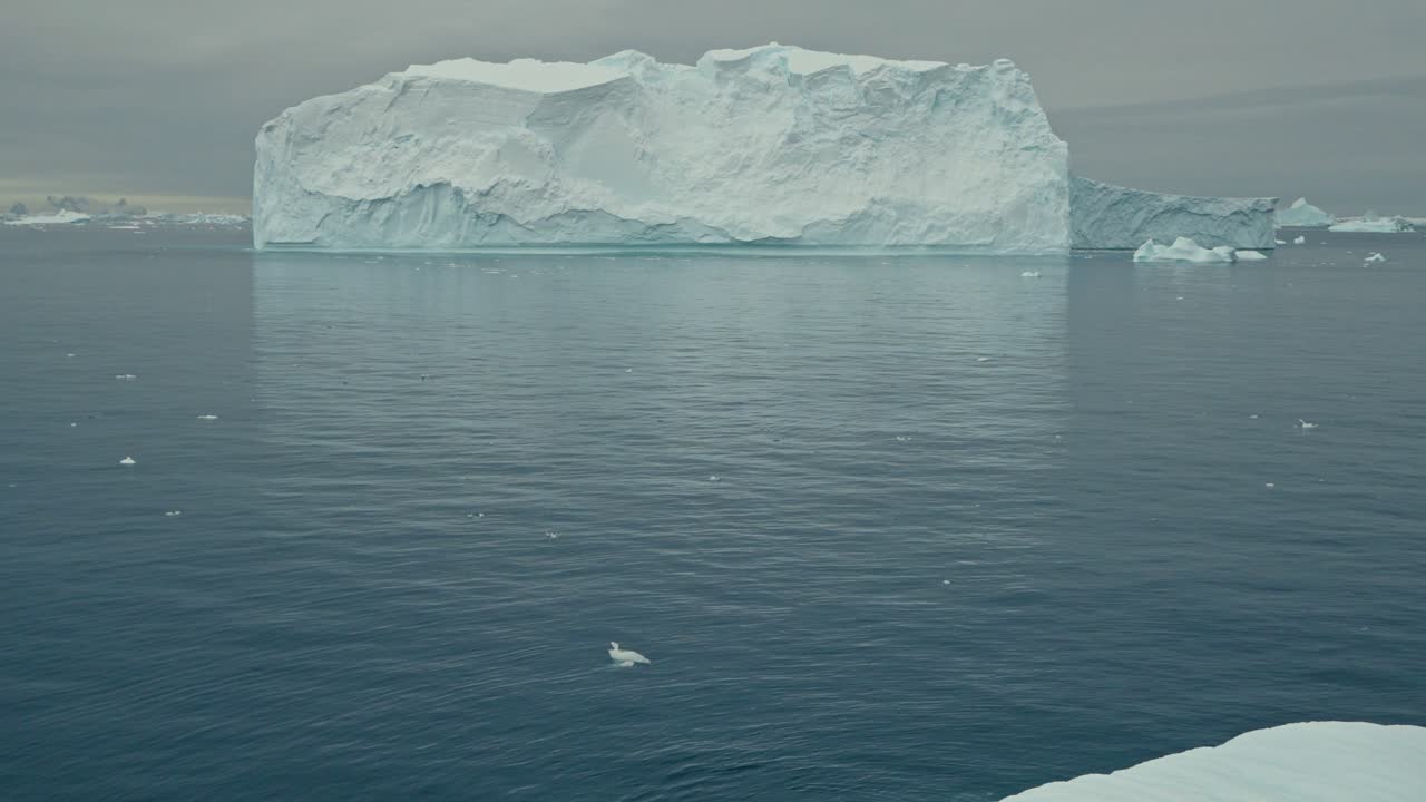 Massive flat-topped tabular iceberg with sheer cliff walls floats in calm grey-blue Antarctic ocean waters surrounded by smaller ice fragments