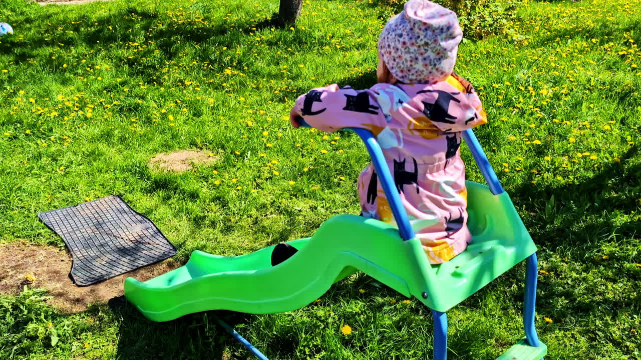 Toddler in warm clothes slides down green plastic slide on sunny day in spring park