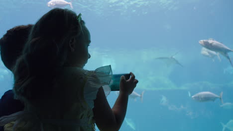 niña tomando fotos de peces en el acuario usando un teléfono inteligente padre e hija fotografiando la vida marina nadando en el tanque con la cámara del teléfono móvil divirtiéndose en el oceanario compartiendo en las redes sociales