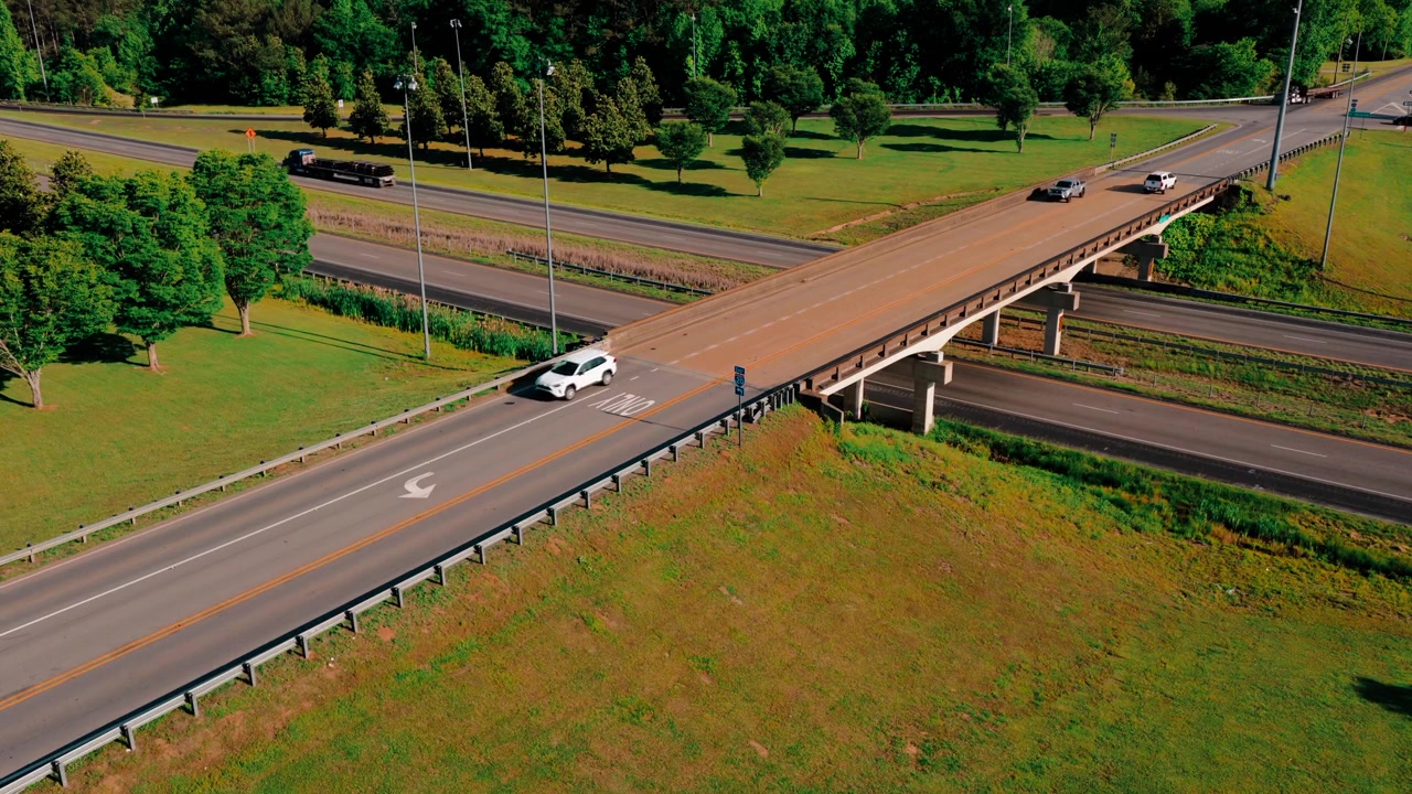 アメリカ合衆国アラバマ州ヘフリンのI-20高速道路で、高架下を通過しながら防水シートで覆われた平床貨物を牽引するセミトラックの空撮ドローンショット。夏の州間高速道路での貨物輸送の様子。