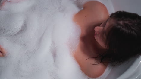 woman covered with fluffy foam lies in bathtub at home. lady enjoys relaxation and spa procedure in bathroom. woman pampers body in hot water