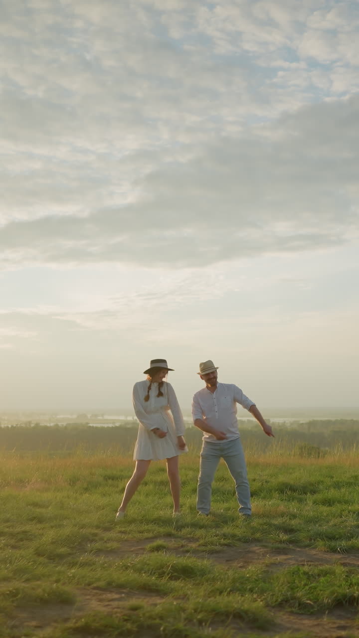 즐거운 부부는 해가 지는 동안 풀이 많은 언덕에서 함께 춤을 추고, 배경에는 조용한 풍경이 있습니다. 색 셔츠와 모자를 입은 남자와 색 드레스를 입은 여자는 손을 놀랍게 움직입니다.