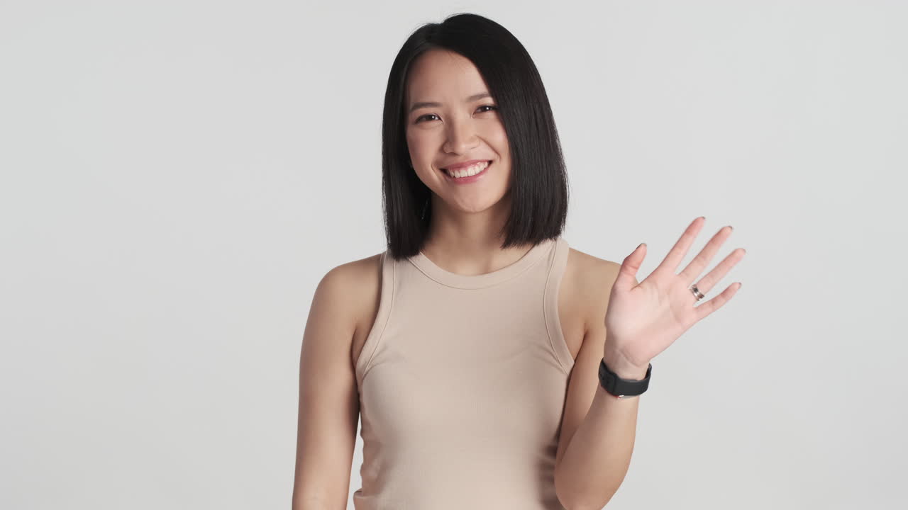 Asian woman smiling and waving hello on camera.