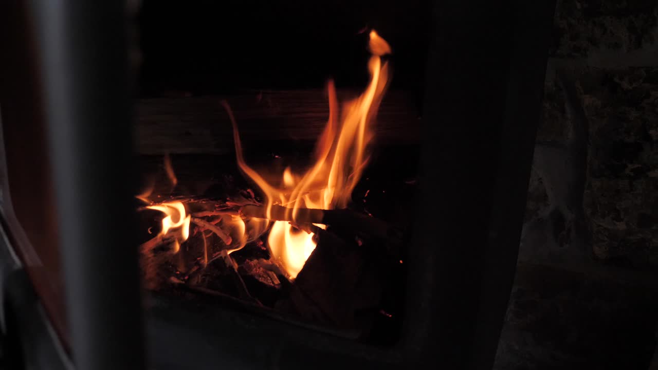 Hand opening metal door of small cabin fireplace as red hot fire burns wood and sticks with slow motion flames flickering with ember and sparks flying up.