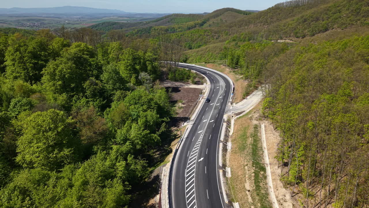 Widok z lotu ptaka przedstawia nowoczesną autostradę asfaltową wijącą się przez gęsty zielony las, symbolizującą infrastrukturę i łączność komunikacyjną.