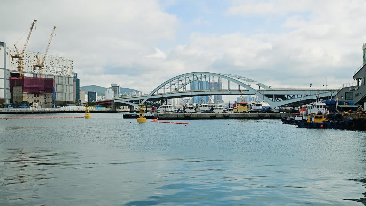 Wide shot of Busan Bridge spanning the harbor, with boats docked below and city buildings including construction sites under a cloudy sky. Urban waterfront view in South Korea.