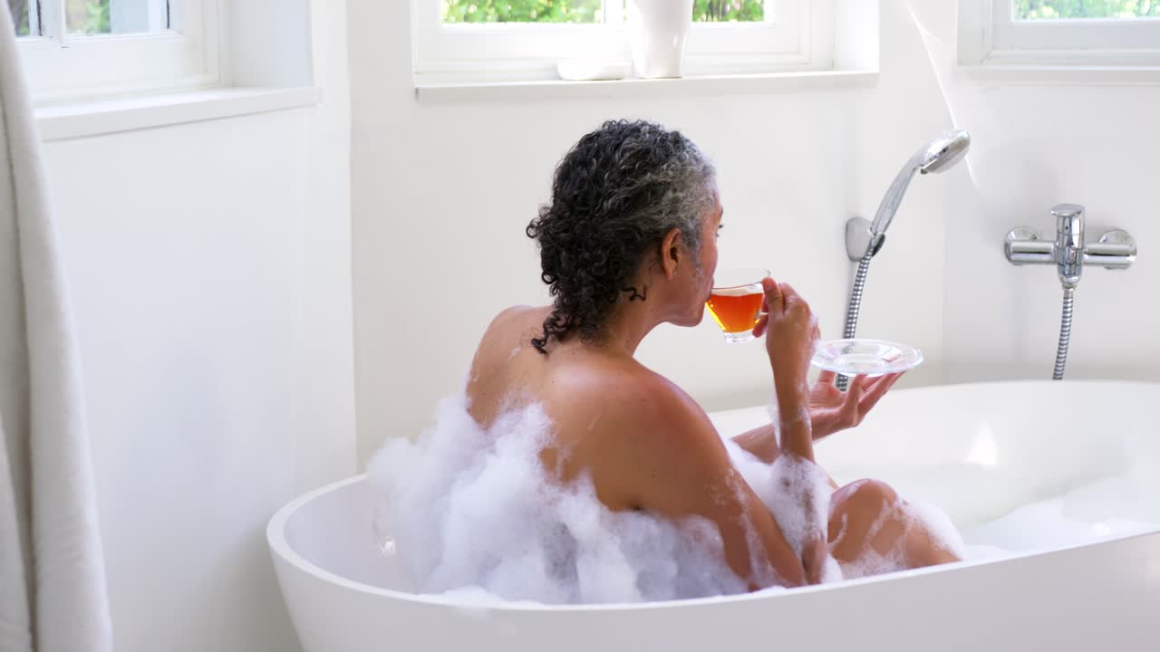 Senior African woman sitting in freestanding bathtub in foam, holding teacup, sipping, relaxing