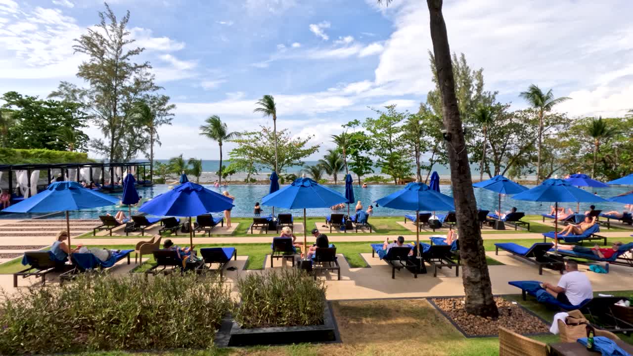 Sunny resort pool area with tourists lounging, blue umbrellas, palm trees, and gentle camera pan