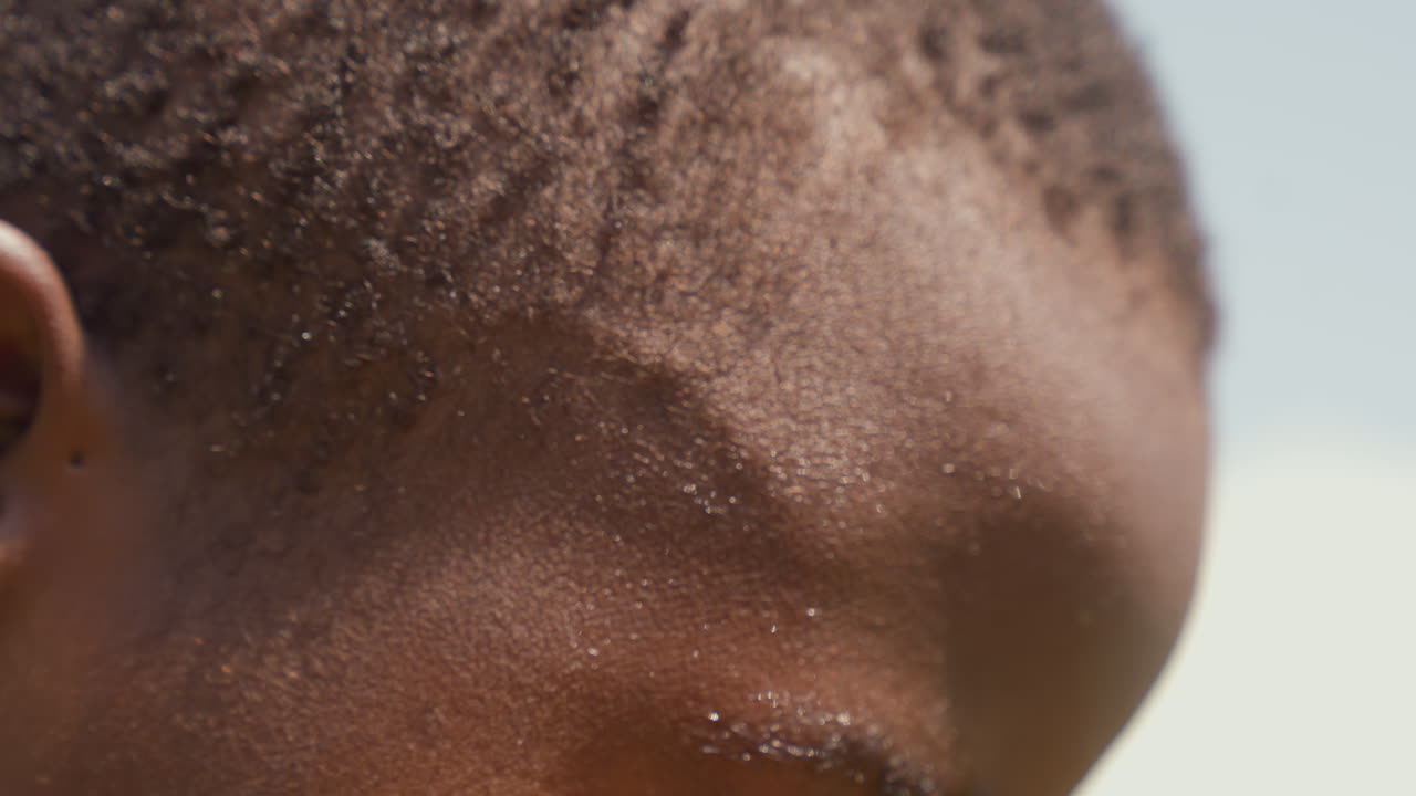 Black boy closeup with sweat, textured scalp and glistening forehead, tight crop on eye and ear, quiet postgame recovery and reflective mood, scenes imply tired trainee, recovering athlete, contender