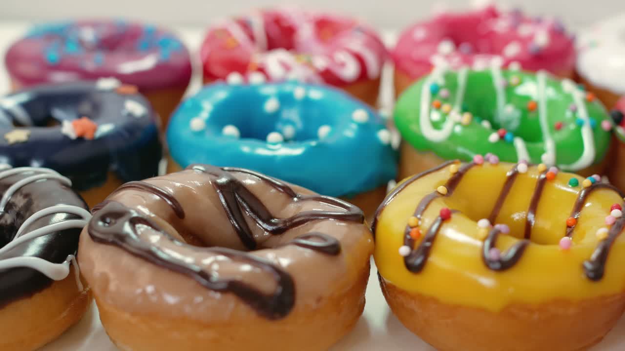 Un gran numero di ciambelle colorate giace su un tavolo bianco in primo piano. Le ciambelle sono decorate con glassa di diversi colori e ricoperte di cioccolato bianco e fondente. Cibo grasso.