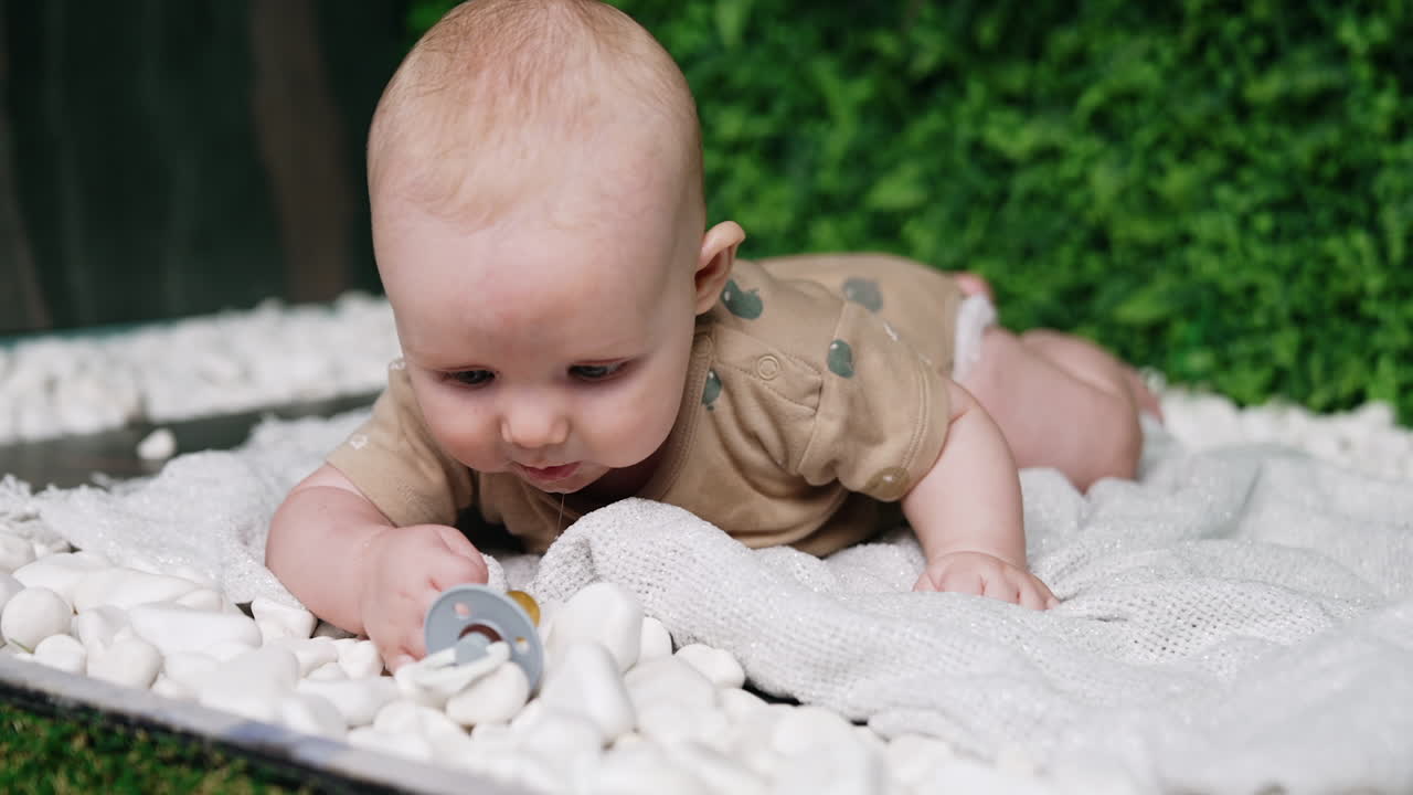 Lovely Caucasian infant lies on white plaid outdoors. Kid lying on the belly looks around and on his pacifier. Close up. Blurred backdrop.