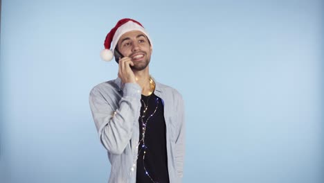 young happy emotional man wearing christmas santa hat and garland on neck standing and talking by mobile phone. man in casual