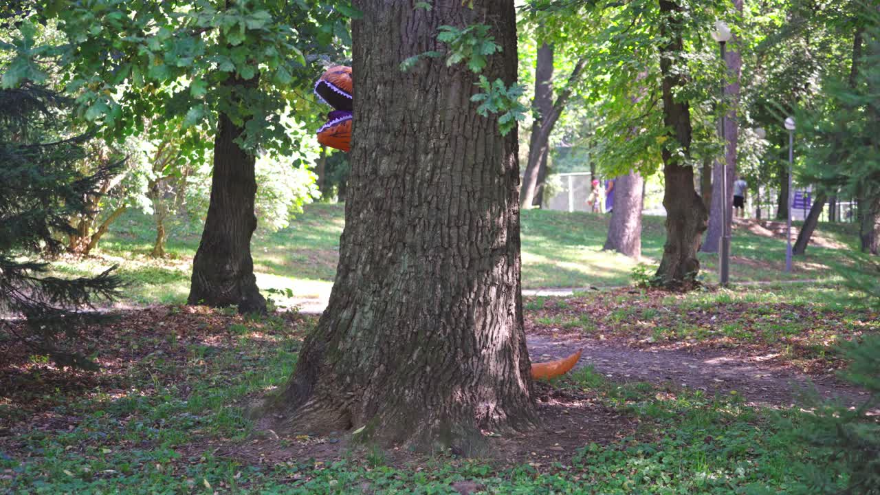 Person in funny suit plays. Dragon blow-up costume hiding behind the tree in the park. Person having fun wearing animal costume outdoors.