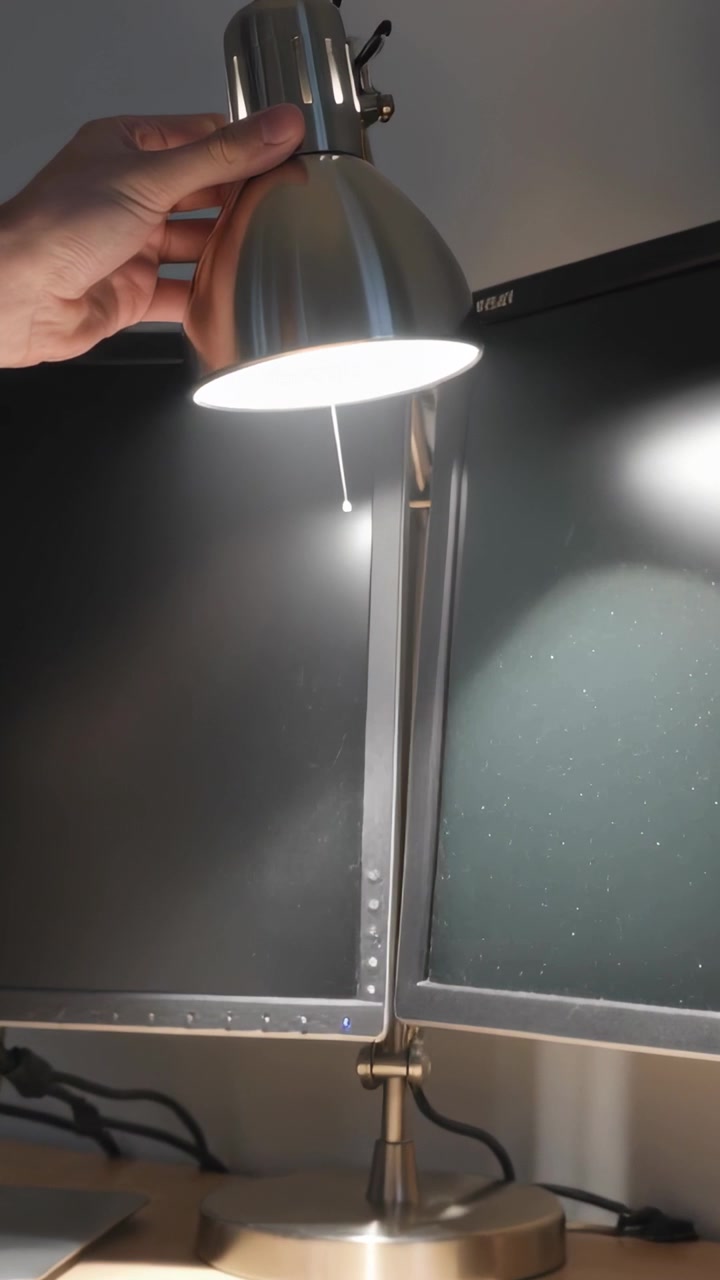 Vidéo verticale: Un bras nu tend la main pour ajuster une lampe métallique sur un bureau près de deux écrans, pour l'éclairage.