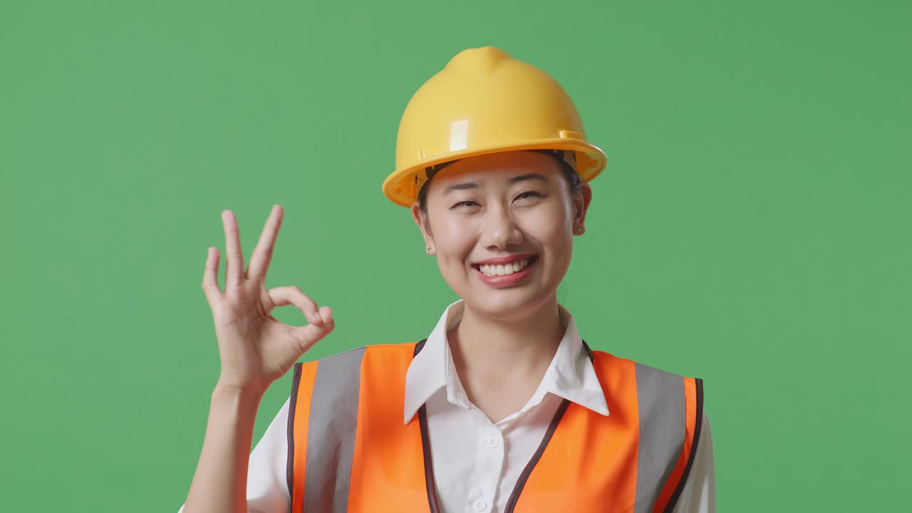 안전 헬을 입은 아시아 여성 엔지니어가 녹색 스크린 배경 스튜디오에 서 있는 동안 미소 짓고 은 몸짓을 카메라에 보여주고 있습니다.