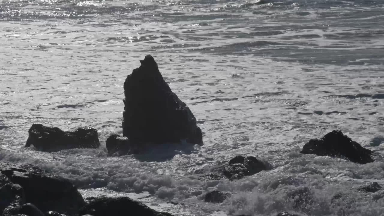 Foamy waves crash powerfully against a single jagged rock formation along Big Sur’s rugged shoreline during daylight.