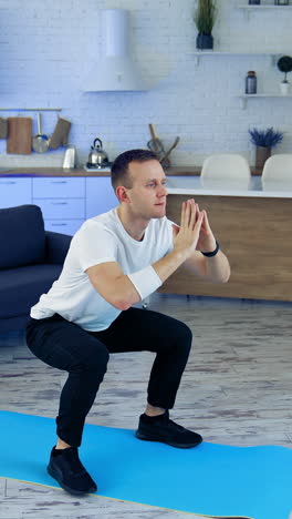 young man exercising on fitness mat at home. active male in white t-shirt and black pants doing his workout on quarantine on kitchen background. sport, fitness and healthy lifestyle at home. vertical video