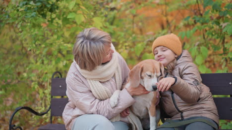 kid holding dog ear playfully while mom watches affectionately, autumn leaves background, beagle seated on bench between them, warm jackets and scarves visible, gentle family bond