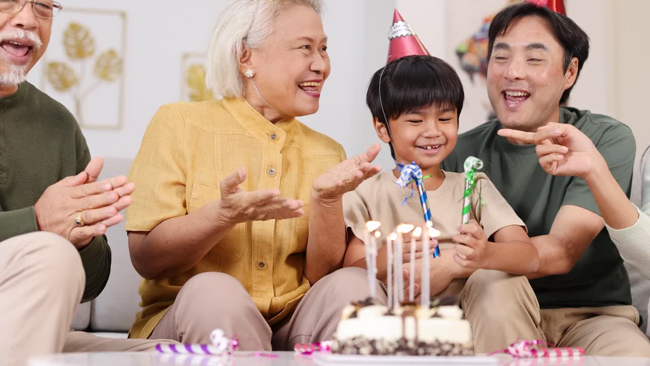 Multigenerational Asian family celebrating birthday with cake, candles, and party hats in cozy home