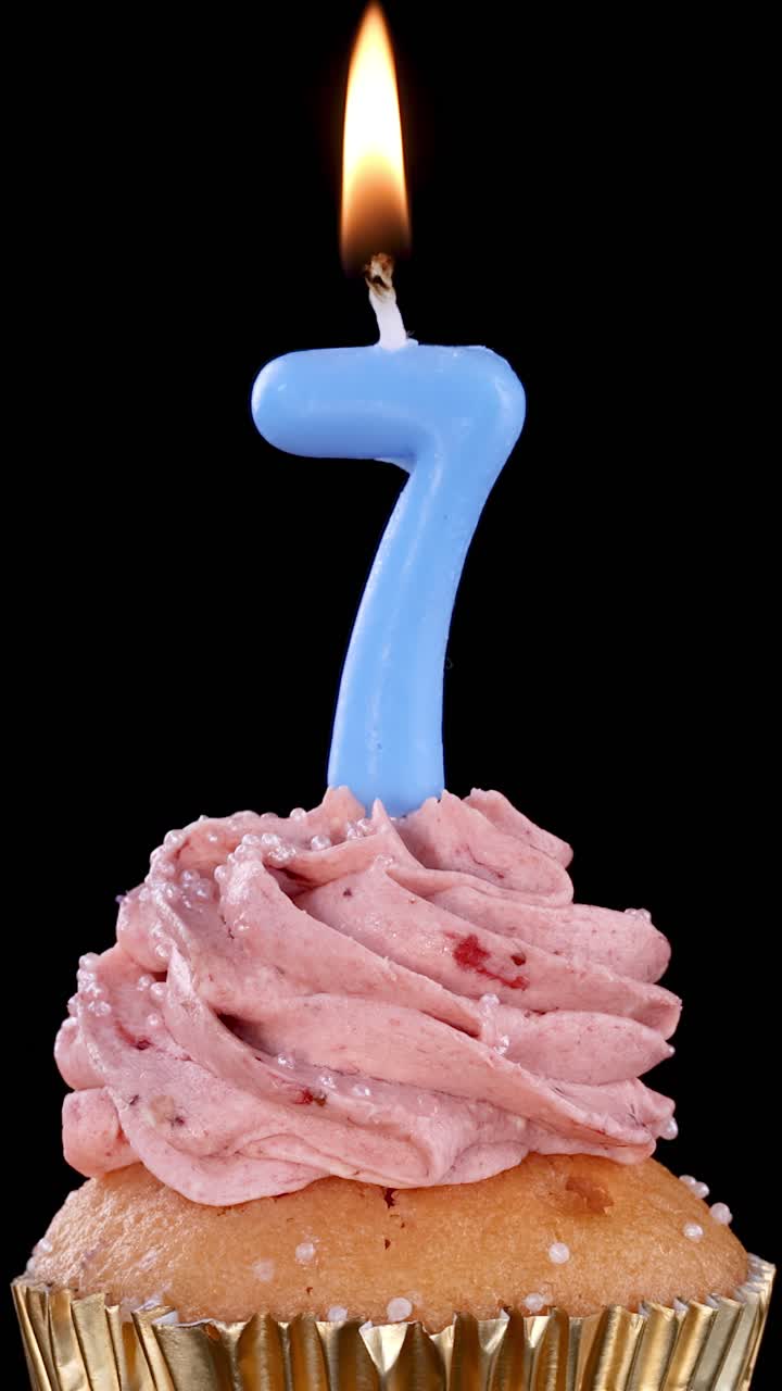 Una candela di compleanno blu a forma di numero sette brucia su un cupcake glassato alla fragola con pirottino dorato, su uno sfondo nero con illuminazione costante e senza movimento della fotocamera