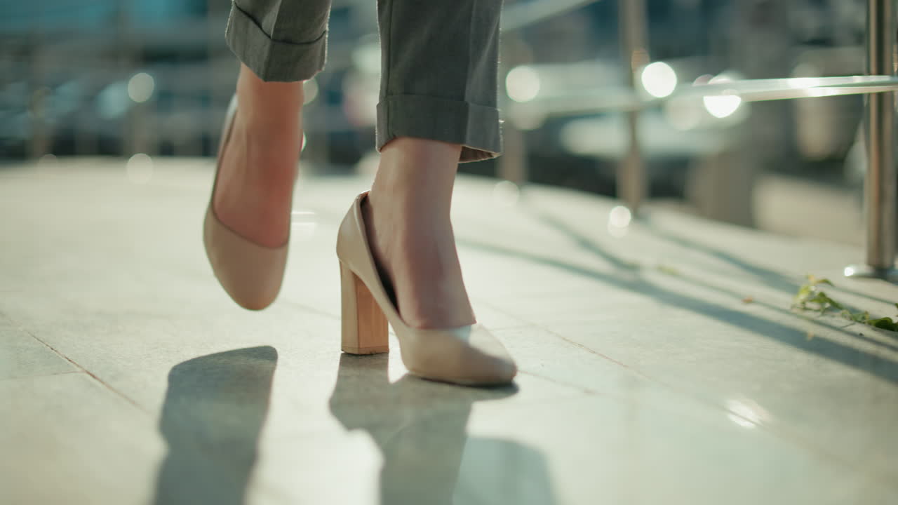Close up leg view of woman wearing heels walking confidently along iron railing with sunlight reflecting on pavement, blurred parked cars and pedestrians in background