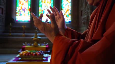 a close-up of a person in orange robes with hands raised in prayer or blessing, set against a backdrop of colorful stained glass windows and offerings
